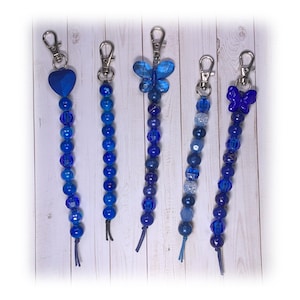 May include: Five blue beaded keychains with different charms. The charms include a heart, a butterfly, and a bow. The beads are various shades of blue.