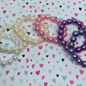 May include: Three pearl bracelets in white, pink, and purple. The bracelets are made of faux pearls and are stacked on top of each other.