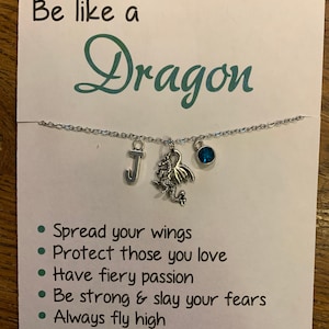 May include: A white card with the text "Be like a Dragon" and a list of inspirational phrases: "Spread your wings", "Protect those you love", "Have fiery passion", "Be strong & slay your fears", "Always fly high", "Rise up and soar". A silver necklace with a dragon charm, a letter "J" charm, and a blue gemstone charm is displayed on the card.