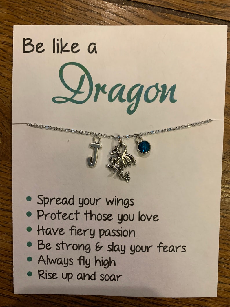 May include: A white card with the text "Be like a Dragon" and a list of inspirational phrases: "Spread your wings", "Protect those you love", "Have fiery passion", "Be strong & slay your fears", "Always fly high", "Rise up and soar". A silver necklace with a dragon charm, a letter "J" charm, and a blue gemstone charm is displayed on the card.