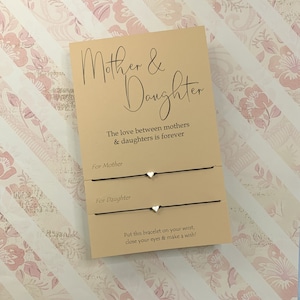 May include: A card with the text "Mother & Daughter" and the phrase "The love between mothers & daughters is forever". Two black string bracelets with silver heart charms are attached to the card. The card also says "Put this bracelet on your wrist, close your eyes & make a wish!"