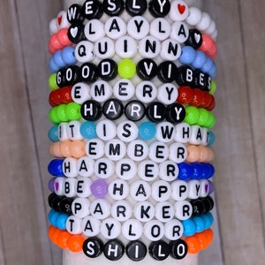 May include: A stack of colorful beaded bracelets with names spelled out in black letters. The names include Wesley, Layla, Quinn, Good Vibes, Emery, Harley, It Is What, Ember, Harper, Be Happy, Parker, Taylor, and Shiloh.