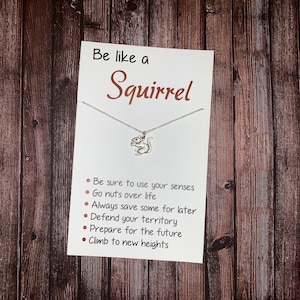 May include: A white card with the text "Be like a Squirrel" in brown lettering. A silver necklace with a squirrel charm hangs from the card. The card lists six tips for being like a squirrel: "Be sure to use your senses", "Go nuts over life", "Always save some for later", "Defend your territory", "Prepare for the future", and "Climb to new heights".