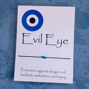 May include: A white card with a blue evil eye charm and the text "Evil Eye" printed in black. The card also includes the text "Protection against danger, evil, bad luck, misfortune, and injury".
