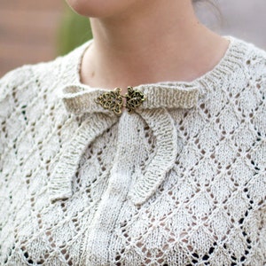 Puede incluir: Un cárdigan de punto blanco con un delicado patrón de encaje y un cierre dorado decorativo en el cuello.