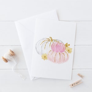 K&ouml;nnte beinhalten: Eine Gru&szlig;karte mit einer Aquarellillustration von drei K&uuml;rbissen in Wei&szlig; und Rosa, akzentuiert mit goldenem Glitzer. Die Karte wird von einem wei&szlig;en Umschlag, einer Garnrolle und zwei kleinen W&auml;scheklammern begleitet.