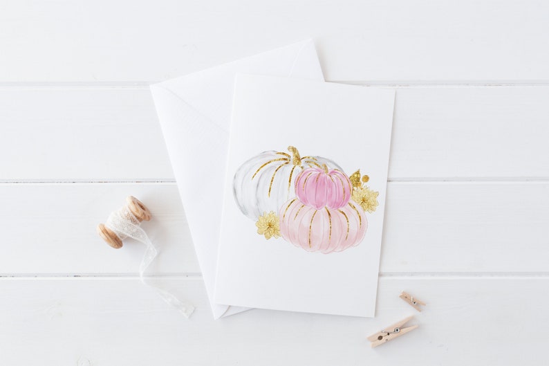 K&ouml;nnte beinhalten: Eine Gru&szlig;karte mit einer Aquarellillustration von drei K&uuml;rbissen in Wei&szlig; und Rosa, akzentuiert mit goldenem Glitzer. Die Karte wird von einem wei&szlig;en Umschlag, einer Garnrolle und zwei kleinen W&auml;scheklammern begleitet.