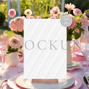 Puede incluir: Un cartel blanco en blanco con una base de madera se encuentra sobre una mesa puesta con platos rosas y blancos, vasos y flores. El cartel es una maqueta para un número de mesa o una tarjeta de lugar.