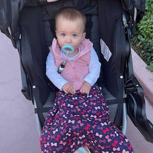 May include: A baby wearing a pink vest and a blue dummy sits in a black pushchair. The pushchair has a navy blue and red bow pattern blanket.