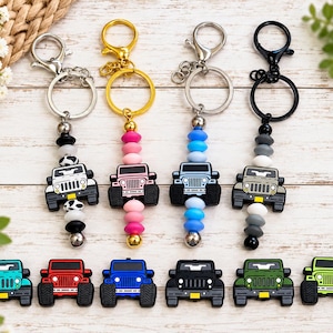 Op de afbeelding: Vier sleutelhangers met siliconen kralen en een kleine SUV-bedel in wit, roze, lichtblauw en grijs, met metalen karabijnsluitingen. Daaronder liggen zes losse SUV-bedels in turquoise, rood, blauw, zwart, donkergroen en limoengroen.