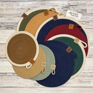 May include: A collection of round, coiled rope trivets in various colors including olive green, mustard yellow, navy blue, burgundy, dark brown, tan, and sage green. Each trivet features a small brown tag with the text "Hand made with love".