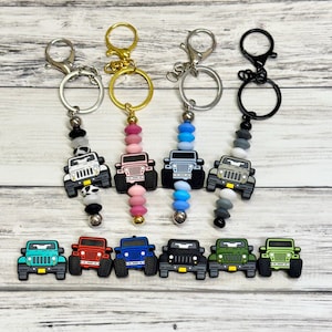 May include: Keychains featuring colorful Jeep charms. The keychains have various bead colors and metal finishes. The charms are in multiple colors, including green, red, blue, and black. The keychains are on a light wood-grain surface.