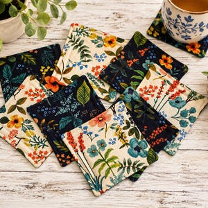 May include: A collection of square fabric coasters featuring floral patterns in cream and dark navy blue backgrounds. The designs include various botanical illustrations of flowers, leaves, and stems in shades of orange, yellow, and blue.