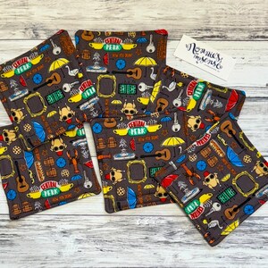 Puede incluir: Un juego de seis posavasos cuadrados de tela con un fondo gris oscuro. Los posavasos presentan un patrón repetido de imágenes icónicas de la serie de televisión Friends, incluyendo el logo de Central Perk, paraguas y guitarras.