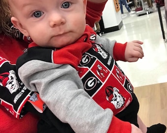 Puede incluir: Un bebé con un mono rojo y negro de la Universidad de Georgia con una camiseta de manga larga gris debajo. El mono tiene un patrón repetido del logotipo de la Universidad de Georgia y una mascota bulldog.
