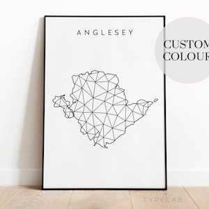 May include: A black and white geometric map of Anglesey, a Welsh island, with the word "Anglesey" printed above the map. The map is framed in a black frame and the background is white. The text "CUSTOM COLOUR" is printed in a circle in the upper right corner of the image.
