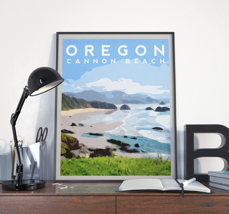 May include: A framed art print featuring a coastal landscape of Cannon Beach, Oregon. The artwork displays a beach scene with ocean waves, rocks, and a blue sky. The print has the text "OREGON CANNON BEACH" in white letters on a blue background.