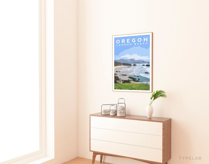 May include: A framed art print featuring a coastal landscape with the text "OREGON CANNON BEACH" in blue. The artwork depicts a beach scene with ocean waves, rocks, and green grass. The print is displayed on a white and wood-tone dresser.