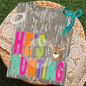 Peut inclure: Un t-shirt &agrave; manches courtes avec un motif camouflage, un graphique de t&ecirc;te de cerf et le texte HE'S HUNTING en lettres rose vif, orange, vert citron et bleu sarcelle. Le t-shirt est orn&eacute; d'un ruban bleu sarcelle et d'un collier de perles.