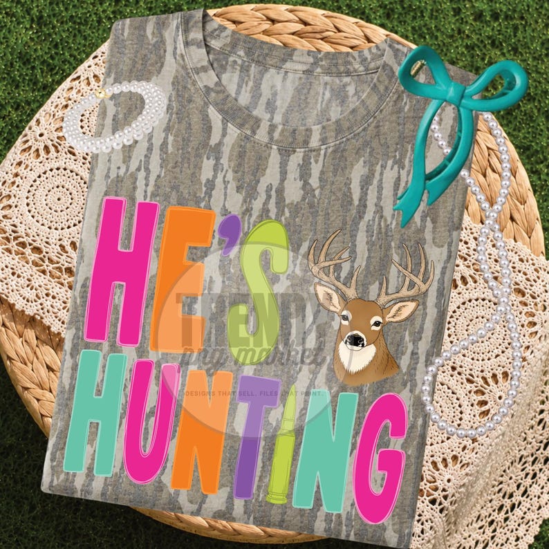 Peut inclure: Un t-shirt &agrave; manches courtes avec un motif camouflage, un graphique de t&ecirc;te de cerf et le texte HE'S HUNTING en lettres rose vif, orange, vert citron et bleu sarcelle. Le t-shirt est orn&eacute; d'un ruban bleu sarcelle et d'un collier de perles.