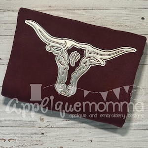 Könnte beinhalten: Ein weinroter Stoff mit einem silbergestickten Longhorn-Schädel-Design. Das Design ist auf dem Stoff zentriert. Der Text "Appliquemomma applique and embroidery designs" befindet sich am unteren Rand des Stoffes.