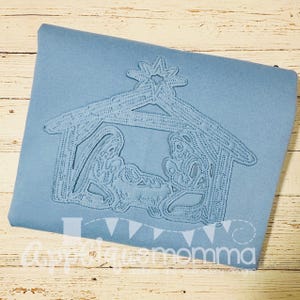 May include: A light blue fabric with an embroidered nativity scene. The design features a star, stable, and figures. The text "AppyMomma" is at the bottom. The design is a light blue colour.