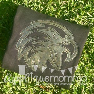 May include: A dark gray shirt featuring a silver glitter eagle design. The eagle is outlined with silver stitching. The shirt is displayed on a bed of green grass. The text "@blaquemom" is visible at the bottom of the shirt.