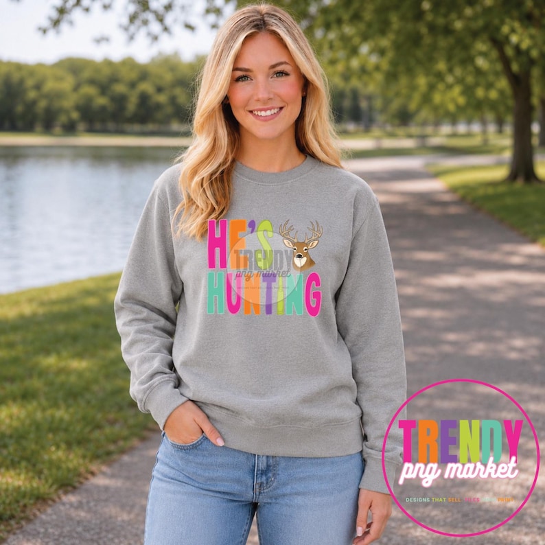 Peut inclure: Un sweat-shirt gris chin&eacute; &agrave; manches longues avec une impression graphique portant le texte HE'S HUNTING en lettres color&eacute;es et une illustration de t&ecirc;te de cerf marron. Le motif inclut un cercle avec le texte TRENDY png market.