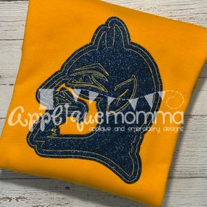 May include: Mustard yellow t-shirt featuring a navy blue glitter applique design. The applique depicts a stylized image of a mascot. The text "Appelque Momma" is printed on the shirt.