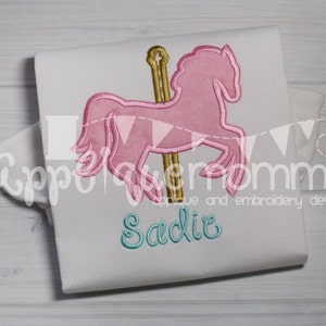 May include: White t-shirt with a pink carousel horse design and gold pole. The name "Sadie" is embroidered in teal script below the horse. The design is an applique and embroidery design.