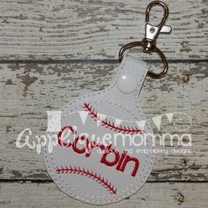 Puede incluir: Llavero de cuero blanco con un diseño de béisbol cosido en rojo y el nombre " Corbin" bordado en rojo. El llavero tiene un clip de metal plateado.