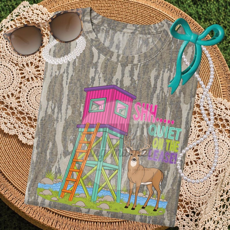 Pu&ograve; includere: Una t-shirt mimetica con la grafica di una torretta da caccia rosa con una scala arancione, un cervo marrone e il testo SHH... QUIET ON THE LEASE! La maglietta &egrave; presentata con occhiali da sole marroni, una collana di perle e un nastro verde acqua.