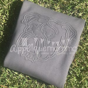 Op de afbeelding: Grijze sweatshirt met een gedetailleerd, gestikt bulldog-ontwerp. Het ontwerp is een donkerdere grijstint en heeft ingewikkelde lijnen. De teksten "appeldem" en "follow" zijn zichtbaar. De sweatshirt ligt op groen gras.