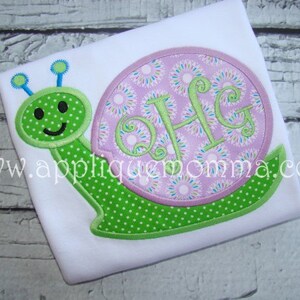 Puede incluir: Un caracol bordado de color verde y rosa con un círculo rosa que contiene el monograma "OHG" en una camiseta blanca.