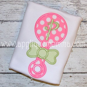 Puede incluir: Un sonajero rosa y blanco con lunares, con un lazo verde y la letra "P" bordada en el sonajero. El sonajero está sobre una camisa blanca.