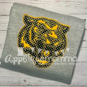 Può includere: Una felpa grigia con un grande disegno a testa di tigre. La testa di tigre è gialla e nera con pois bianchi. Il testo "Appelmomma embroidery designs" è stampato in basso a sinistra.