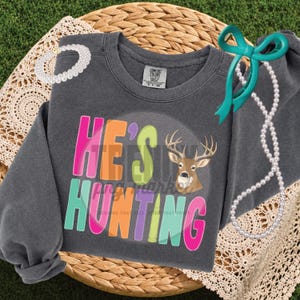 Peut inclure: Un sweat-shirt gris anthracite &agrave; col rond avec le texte color&eacute; HE'S HUNTING et un motif de t&ecirc;te de cerf. Le design comprend des lettres rose vif, orange, vert citron et bleu sarcelle. Un collier de perles blanches et un ruban bleu sarcelle sont pos&eacute;s dessus.