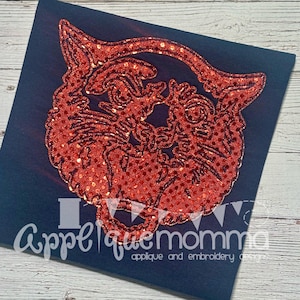 May include: A red glitter applique of a stylized cat face on a navy blue background. The applique is detailed with a textured, sparkling finish. The text "Applique Momma" is at the bottom, with "applique and embroidery designs" below it.