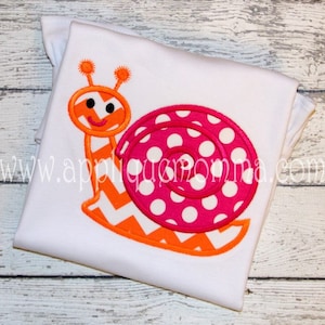 Puede incluir: Una camiseta blanca con un diseño de caracol bordado en naranja y rosa. El caracol tiene un patrón de chevron en su cuerpo y una concha rosa con lunares blancos.