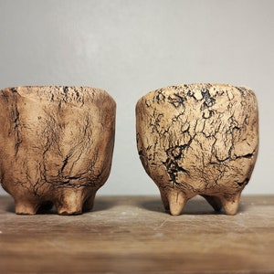 May include: Two small, brown ceramic pots with a crackled glaze and three legs each. The pots are sitting on a wooden surface.