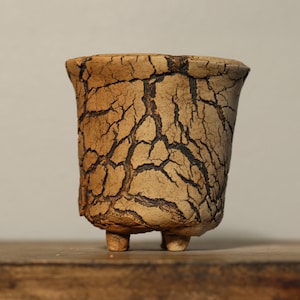 Può includere: Un piccolo vaso in ceramica marrone con una glassa screpolata e tre gambe. Il vaso è appoggiato su una superficie di legno.