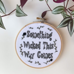 May include: A white fabric embroidery hoop with the phrase "Something Wicked This Way Comes" in dark gray thread. The design includes small floral accents and a wooden hoop frame. A black rose is stitched on the left side.