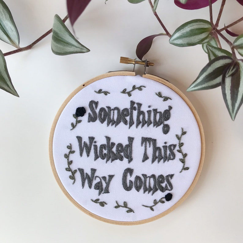 May include: A white fabric embroidery hoop with the phrase "Something Wicked This Way Comes" in dark gray thread. The design includes small floral accents and a wooden hoop frame. A black rose is stitched on the left side.