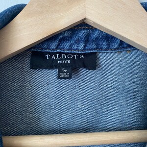 May include: Close-up of a blue denim jacket with the label "TALBOTS PETITE" visible. The jacket is hanging on a wooden hanger. The label also indicates the jacket is size SP and made in Vietnam.