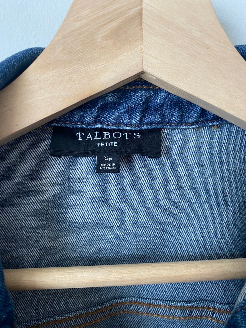 May include: Close-up of a blue denim jacket with the label "TALBOTS PETITE" visible. The jacket is hanging on a wooden hanger. The label also indicates the jacket is size SP and made in Vietnam.