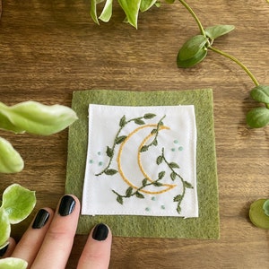 Peut inclure: Un patch brodé carré avec un croissant de lune doré et des vignes vertes sur un fond blanc, posé sur un carré de feutre vert. De petites perles bleues entourent le motif. Le patch est présenté sur une surface en bois.