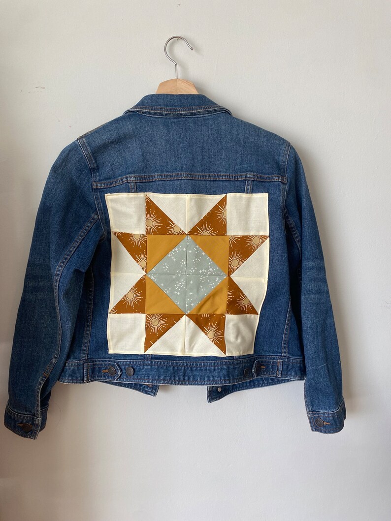 May include: A blue denim jacket featuring a patchwork star design on the back. The star is made from cream, brown, and gold fabric. The centre of the star is a light blue square with white starburst patterns. The jacket hangs on a wooden hanger.