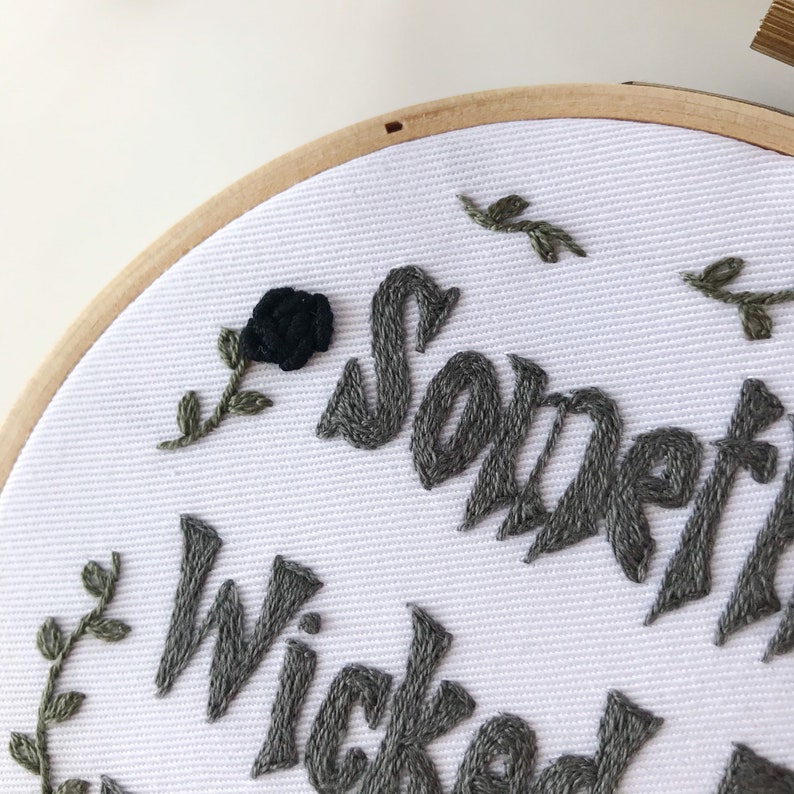 May include: Embroidered hoop art with the words "Something Wicked" in gray thread. A black rose and green leaves are also embroidered on the white fabric. The wooden hoop is visible.