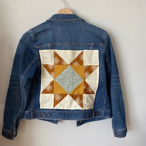 May include: A blue denim jacket with a patchwork star design on the back. The star is made from cream, mustard yellow, and light blue fabrics, with sunburst details. The jacket is suspended from a wooden hanger.