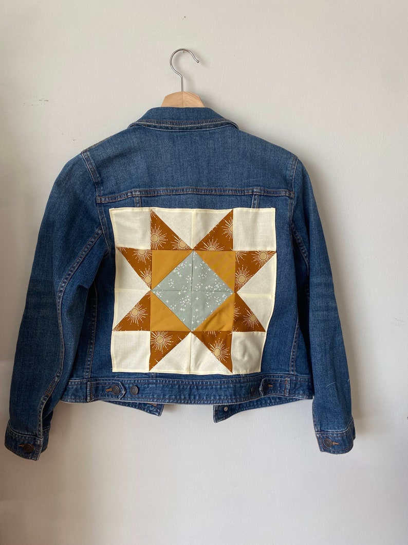 May include: A blue denim jacket with a patchwork star design on the back. The star is made from cream, mustard yellow, and light blue fabrics, with sunburst details. The jacket is suspended from a wooden hanger.